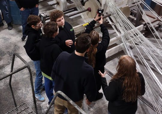 A group of young people on a Bedford factory tour observes fiberglass machinery with interest.