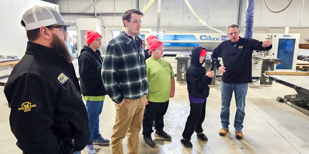 A group of students stand bright hats listen to a Bedford guide pointing at machinery.